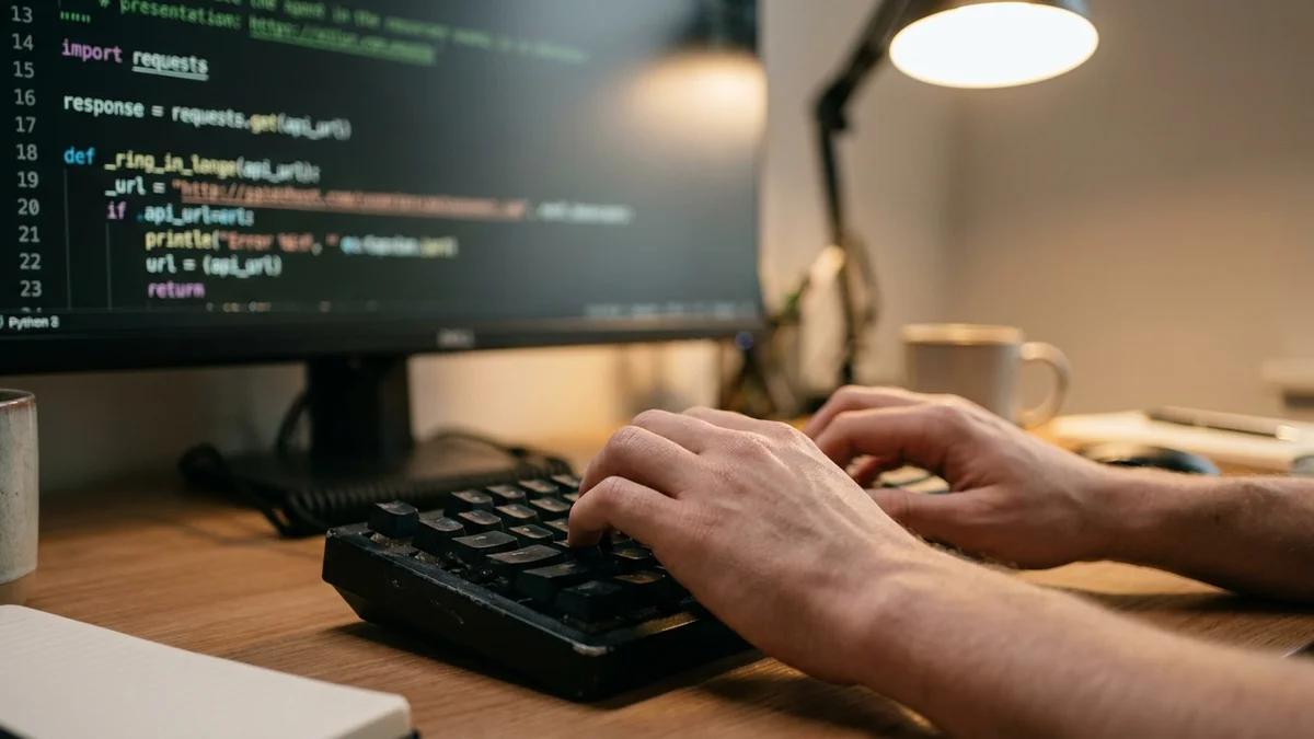 Close-up of hands typing on a mechanical keyboard with Python API code on the monitor behind