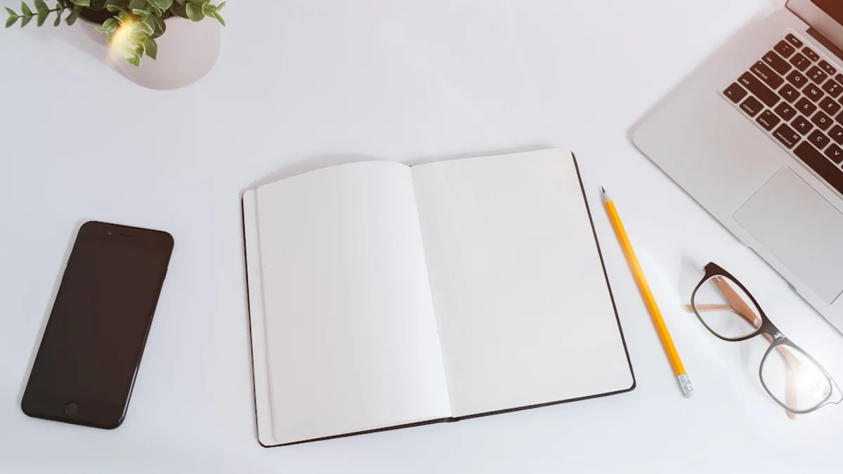 An open empty notebook on a white desk next to an iPhone and a MacBook